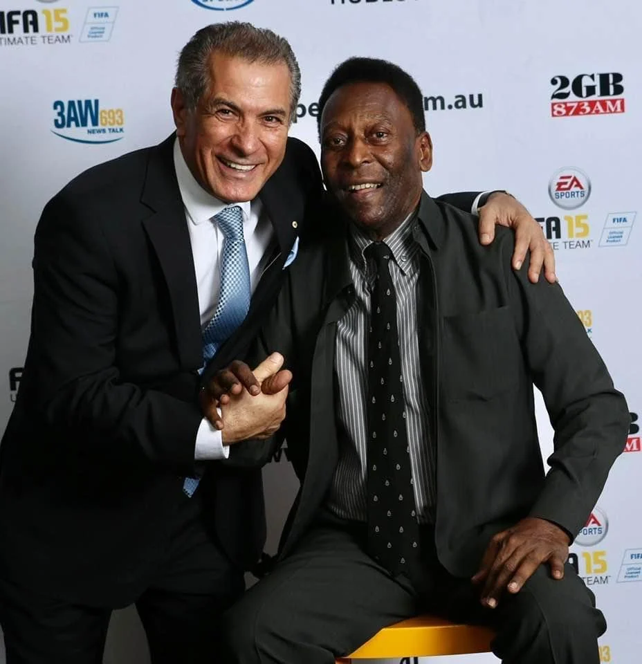 A smiling man in a dark suit leans in with his arm around Pelé, who is seated in a black suit, in front of a sponsors’ media backdrop at a football function.
