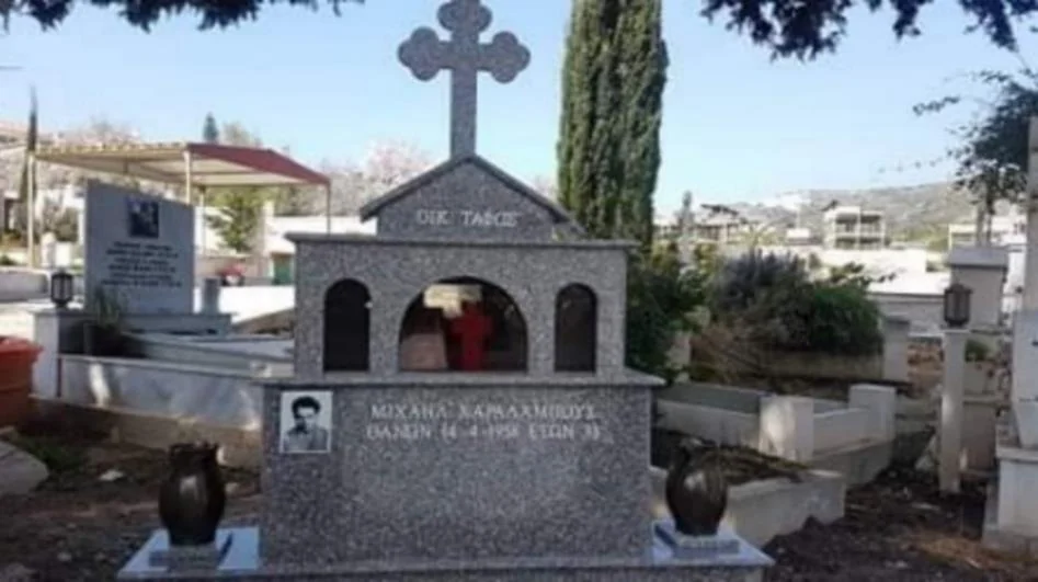 A new dark granite memorial with a stone cross and an inscribed plaque, standing among older white graves in a sunlit cemetery with cypress trees and hillside houses behind.
