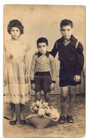 A worn sepia photograph of three children standing side by side, the youngest in shorts between a girl in a pale dress and an older boy, a basket of flowers at their feet.