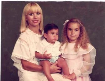 A studio portrait in colour: a woman with long blonde hair in a white lace dress holds a toddler in white on her lap, with a young girl in a pale pink dress beside them.