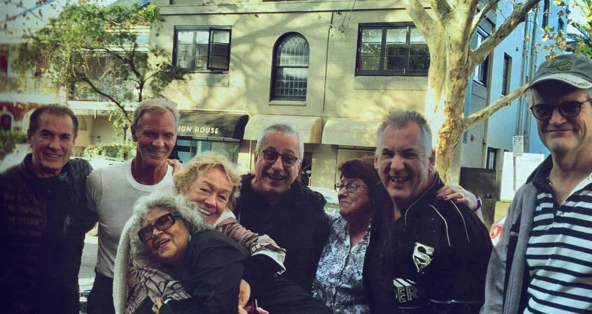 A group of people standing close together outdoors in front of a building, smiling, arms around one another.