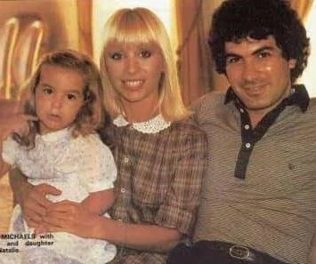 A 1980s colour photograph indoors: a man with dark curly hair beside a woman with blonde hair who is holding a small girl in a white dress.