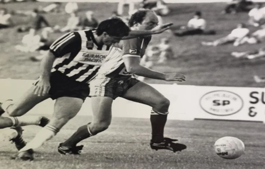 A monochrome match photograph: a player in a black-and-white striped shirt turns away from a defender in a light kit, the ball on the grass beside them and a sparse crowd behind.