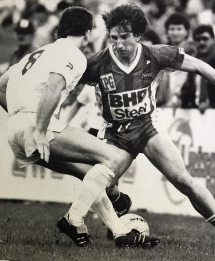 A monochrome match photograph: a player in a dark BHP Steel shirt takes the ball past a defender in white, with spectators behind the advertising boards.
