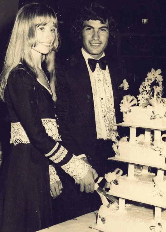 A monochrome photograph of a young couple behind a tall tiered wedding cake; she wears a dark gown with lace cuffs, he wears a dinner jacket and bow tie.