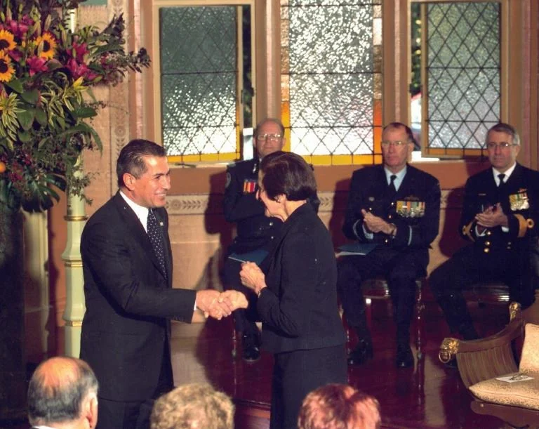 In a formal reception room with leaded windows and a tall floral arrangement, a man in a dark suit shakes hands with a woman while uniformed officers and guests look on.