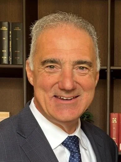 A headshot of Paul Nicolaou, smiling, with grey hair, wearing a dark suit and a blue spotted tie, in front of bookshelves.